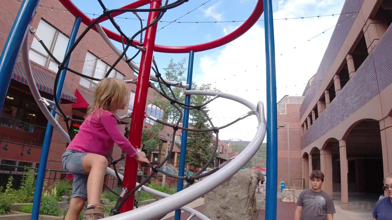 At 0:00 in Winter Park, Colorado, a young girl in a pink top climbs a red and blue rope play structure with brick buildings and hills behind her.