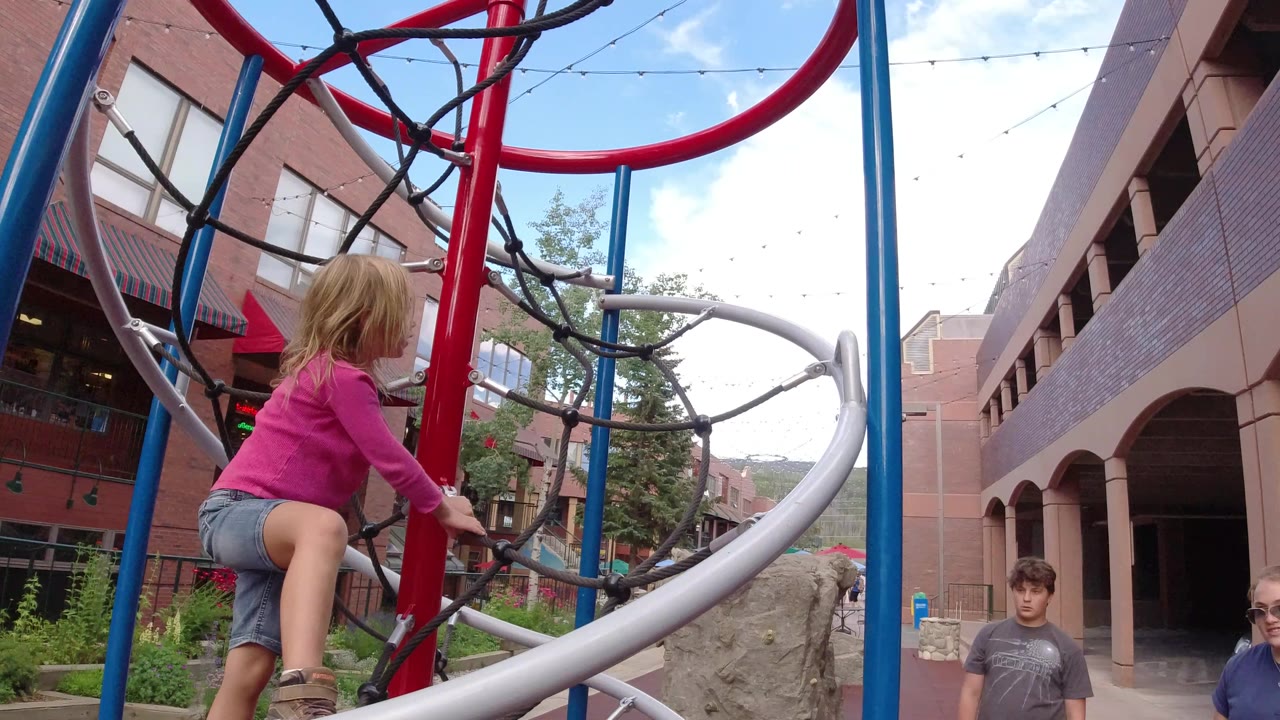 At 0:01 in Winter Park, Colorado, the girl holds the red pole on the play structure while a young man and a woman in sunglasses appear near the walkway.