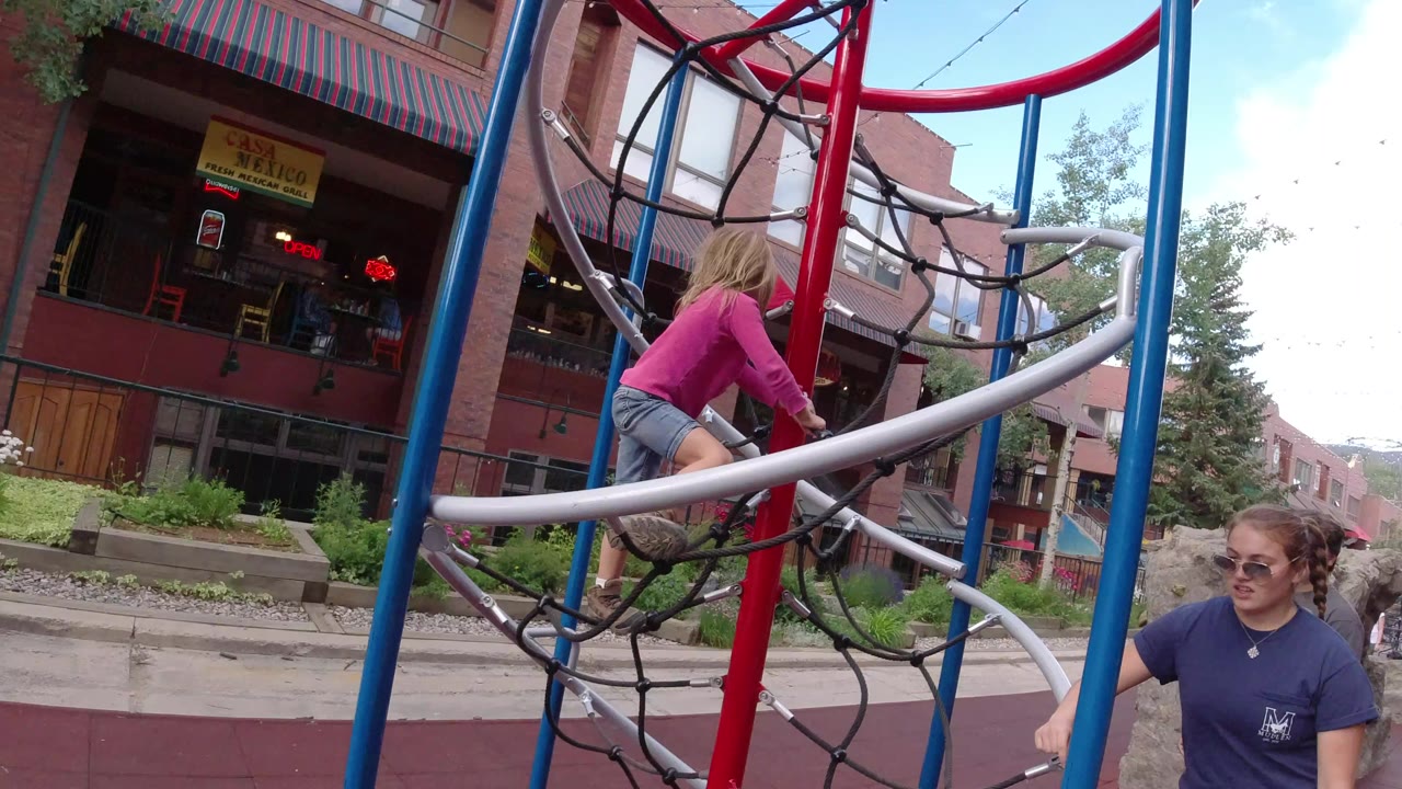 Child climbs a rope net on a playground while an adult stands nearby.
