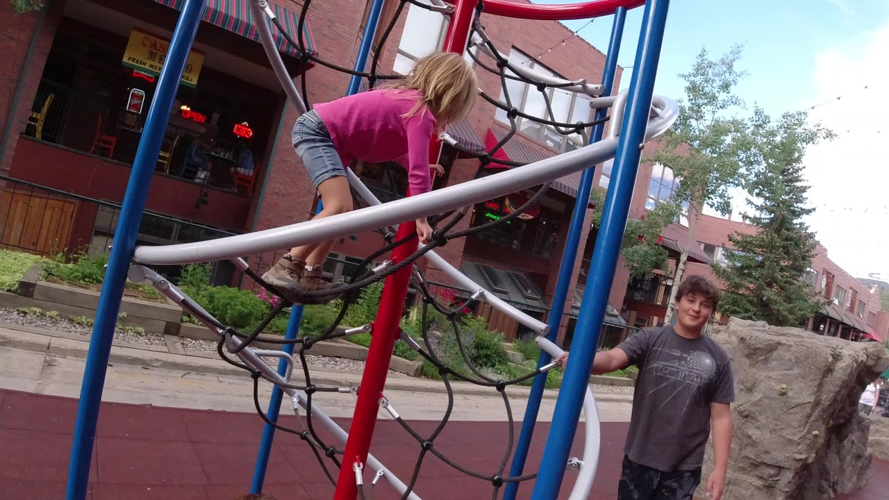 In Winter Park, Colorado, the girl shifts forward on the rope climbing structure while the adult man remains visible beside the play area and storefronts sit behind them.