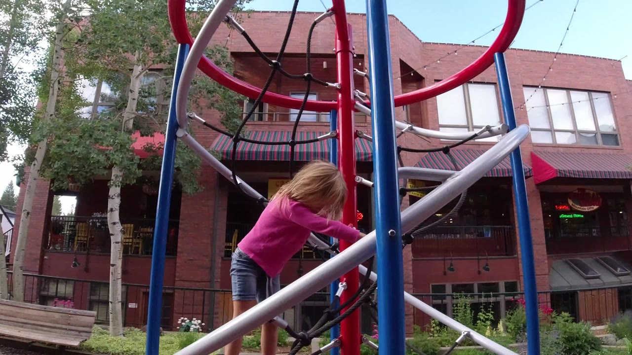 Child in a pink shirt climbing a rope jungle gym near brick buildings.