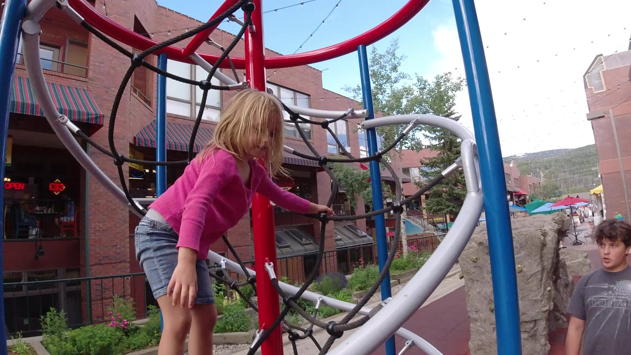 Child climbing a playground rope structure beside a person near brick buildings.