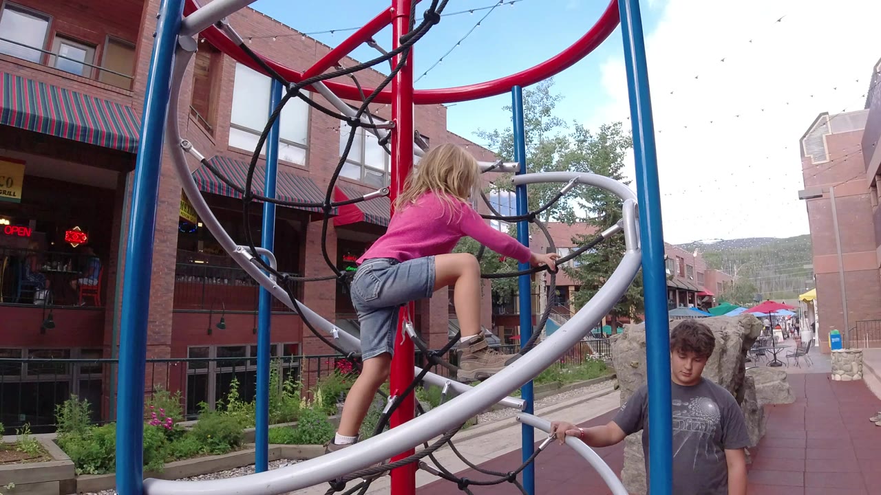 Blonde child climbing playground bars beside brick storefronts.