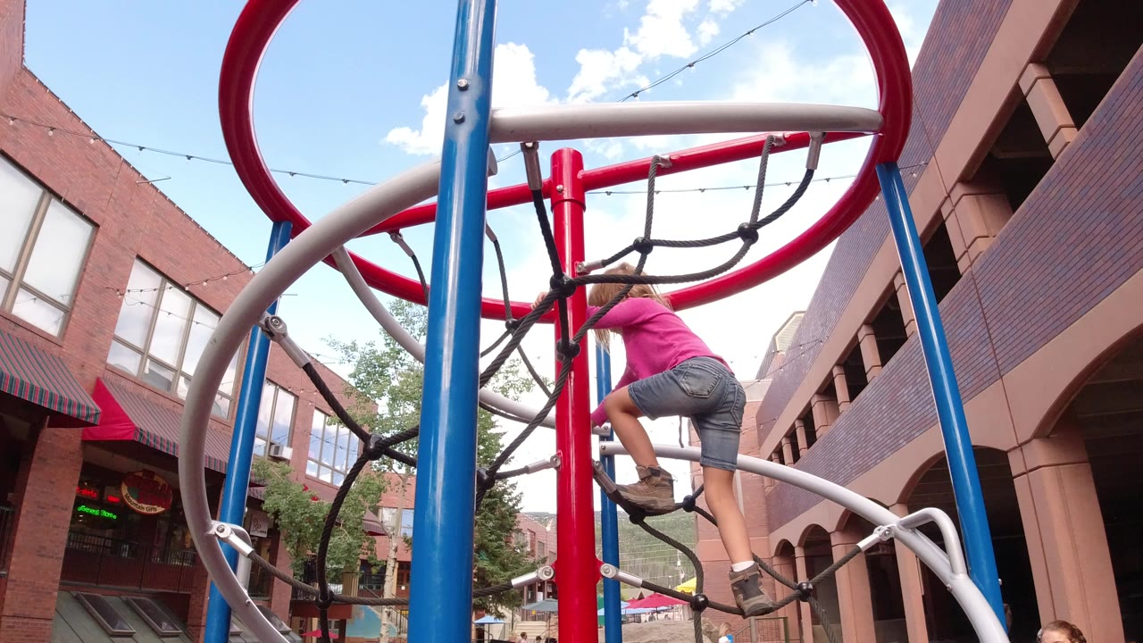 Child climbing a colorful rope playground structure beside brick buildings.