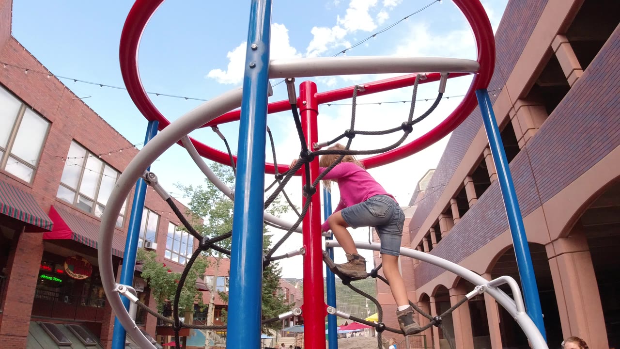 The child balances on the rope net within the colorful playground frame in Winter Park, Colorado, while brick storefront windows fill the background.