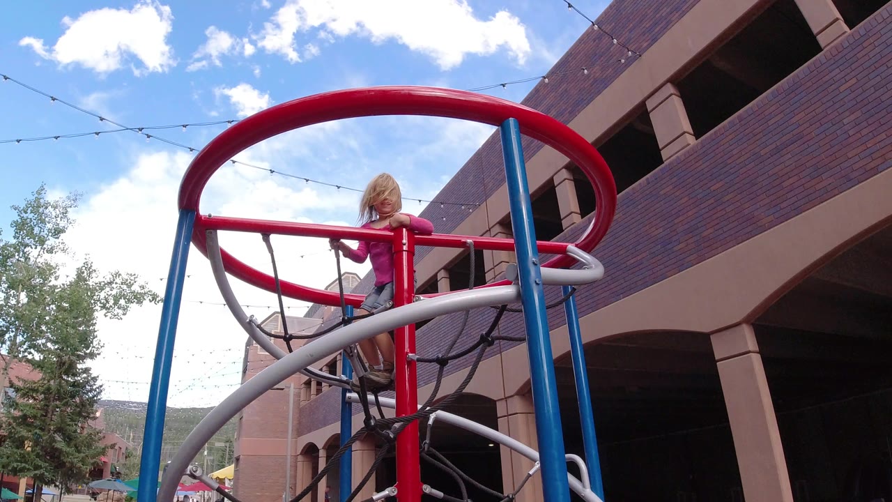 The girl holds the red rail of the jungle gym while standing above black rope netting, with the brick facade and sky still filling the background in Winter Park, Colorado.