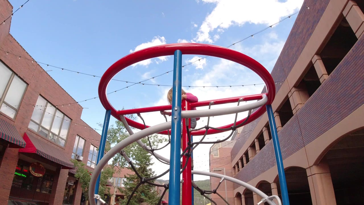 A child in a pink top is partly obscured by a red circular climbing rail and blue posts in Winter Park, Colorado, with brick buildings and sky behind.