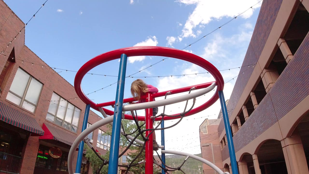 A child in a pink top leans over a red playground rail in Winter Park, Colorado, with brick buildings, string lights, blue sky, and clouds behind the structure.