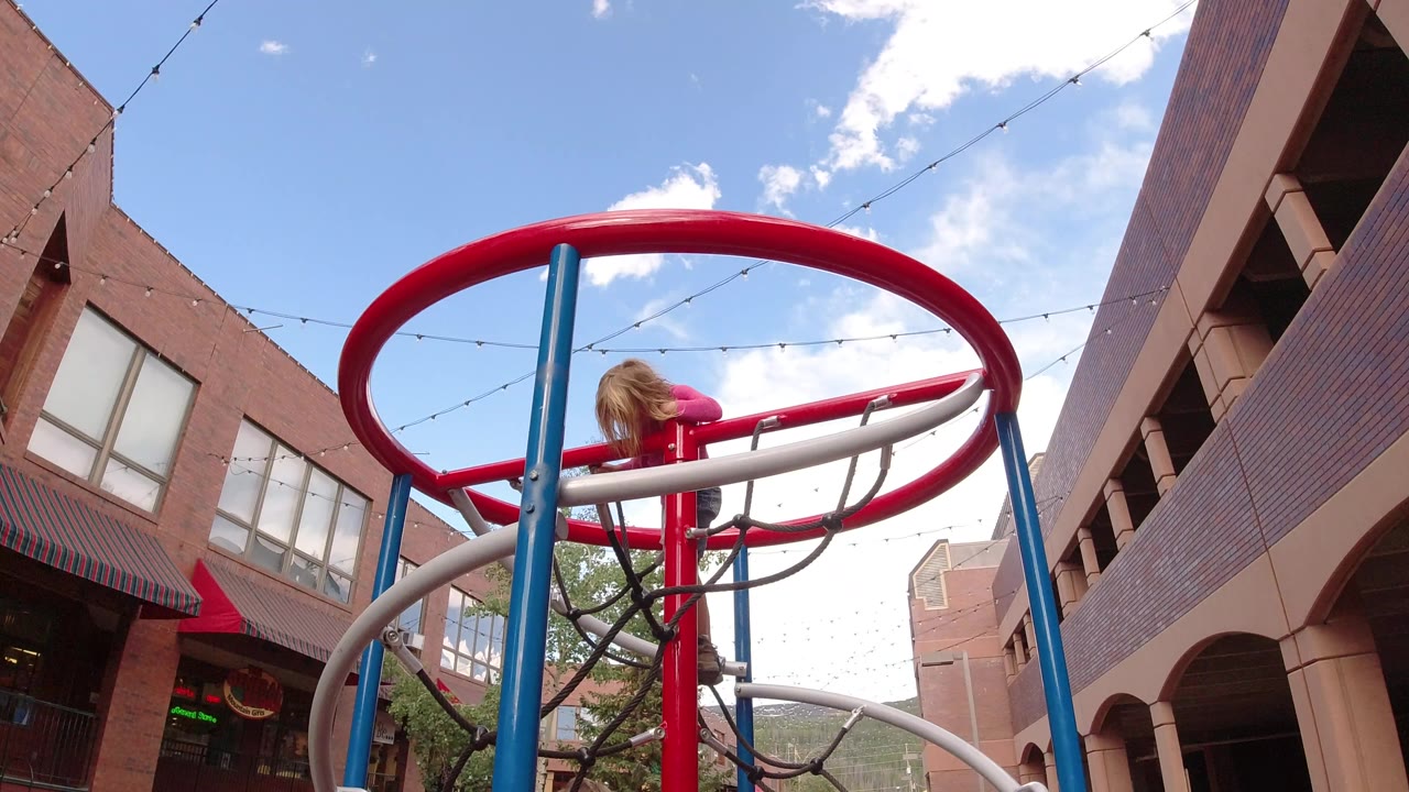 The child remains near the left side of the red circular rail in Winter Park, Colorado, with the blue pole centered and brick storefront-style buildings framing the sky.