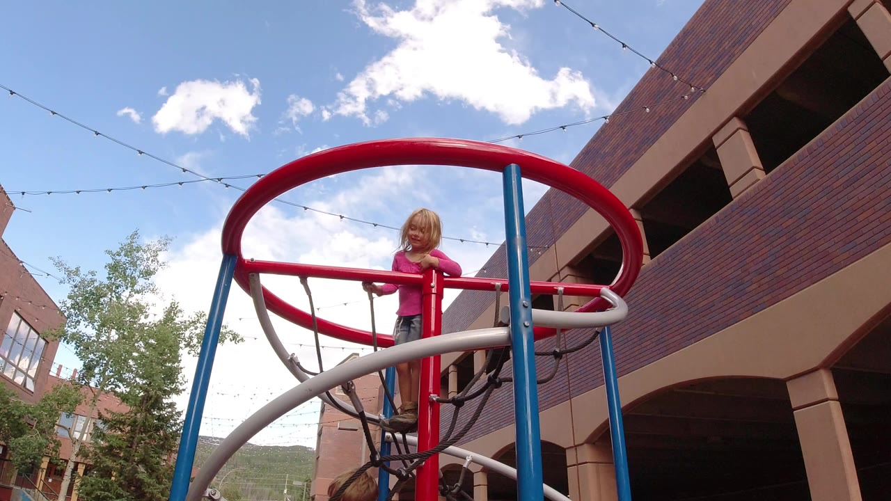 A young girl stands on red playground rails in Winter Park, Colorado, with blue posts, white curved bars, brick buildings, trees, string lights, and clouds behind her.