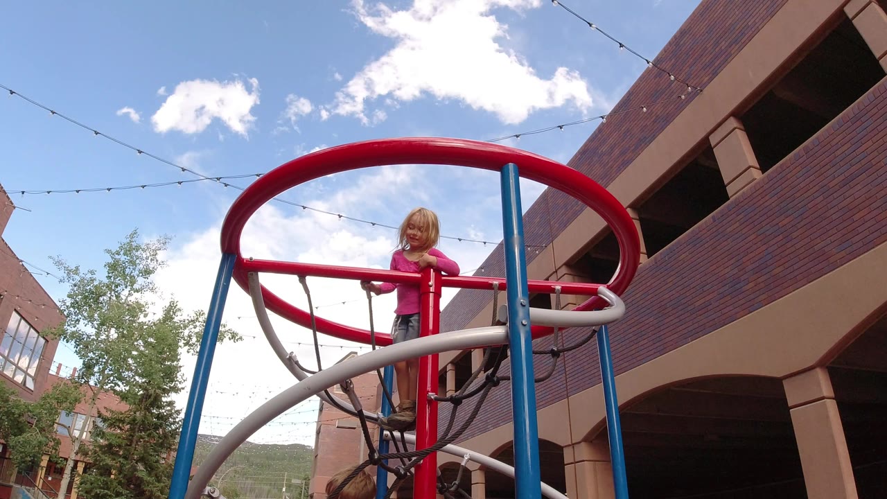 The girl remains centered on the red circular playground frame in Winter Park, Colorado, with a brick building rising at right and string lights crossing the sky.