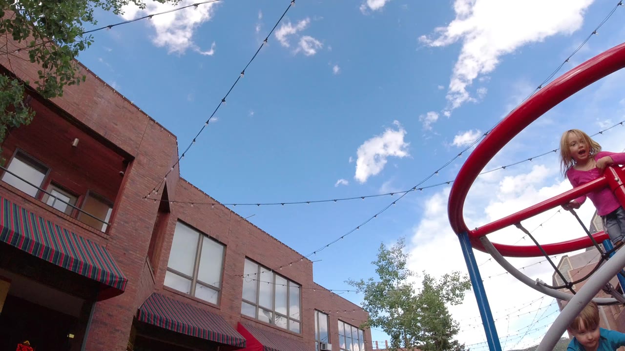 A child in a pink shirt climbs a red playground frame in Winter Park, Colorado, with brick buildings, string lights, clouds, and a second child partly visible below.