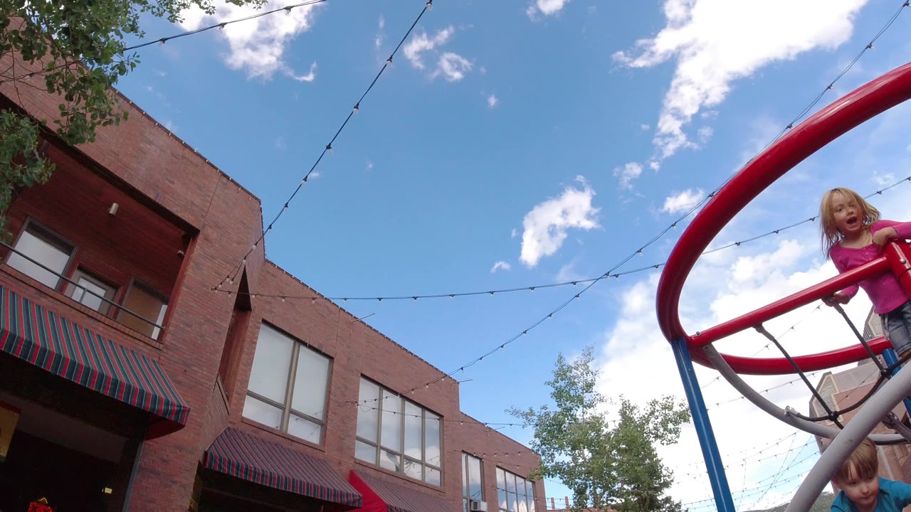 A child in pink leans over the red jungle gym in Winter Park, Colorado, while another child appears beneath the bars against brick buildings and a bright cloudy sky.
