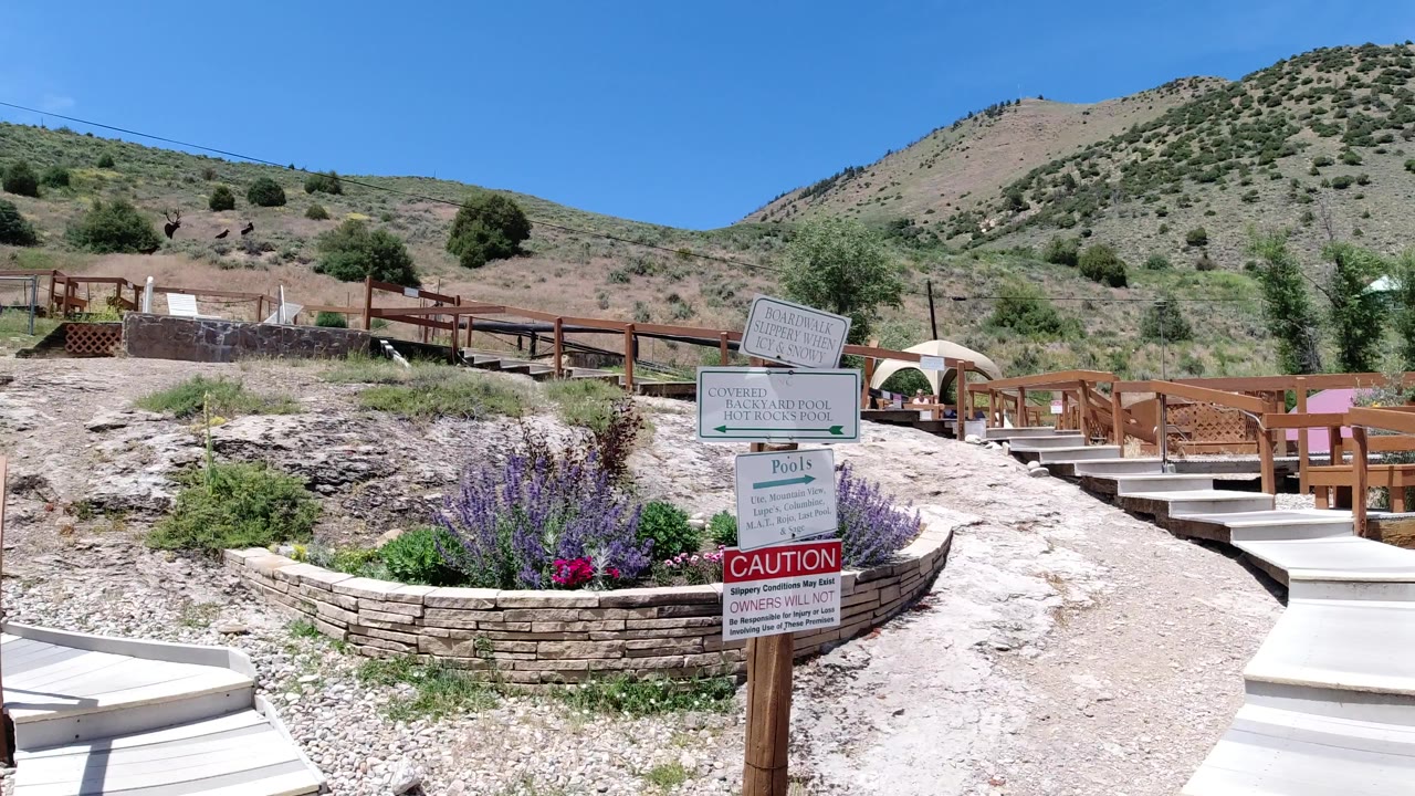 Winter Park, Colorado frame with the signpost closer to center, a gravel path curving past wood railings, planted flowers, and scrub-covered hillside under clear blue sky with no visible people.