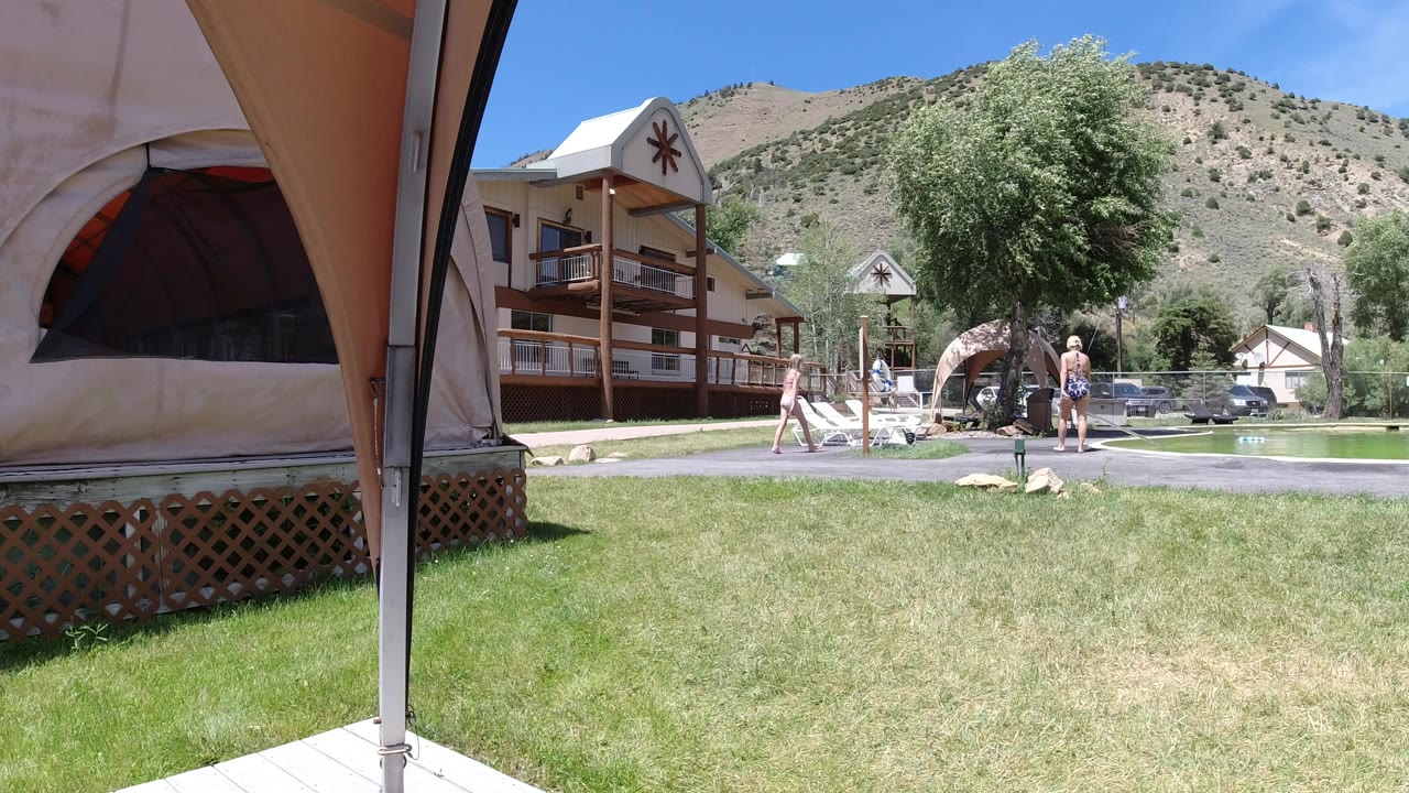 Winter Park poolside frame with a broad grass foreground, people moving around lounge chairs, lodge balconies, leafy trees, and a sunlit hillside behind them.