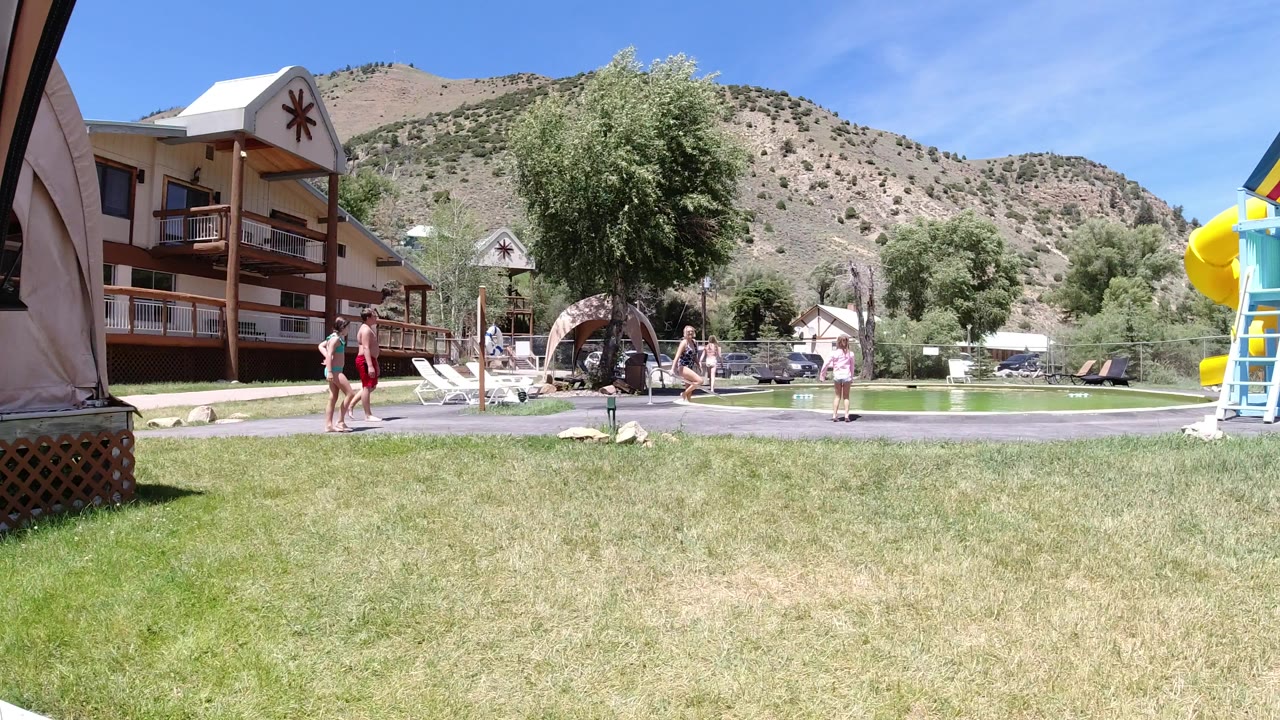 Adults and children move across a sunny Winter Park playground area, with lounge chairs, trees, a lodge-style building, and a hillside behind the play space.