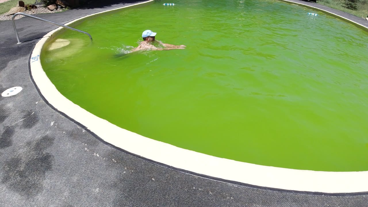 Man in a light cap swimming in a bright green outdoor pool.