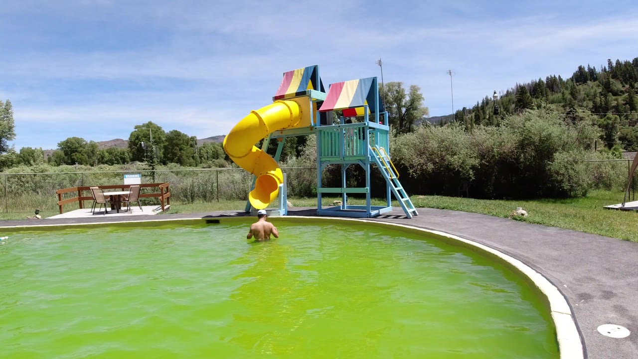 A man seen from behind stands in a bright green pool in Winter Park, Colorado, facing a yellow tube waterslide and blue play structure under a clear sky.