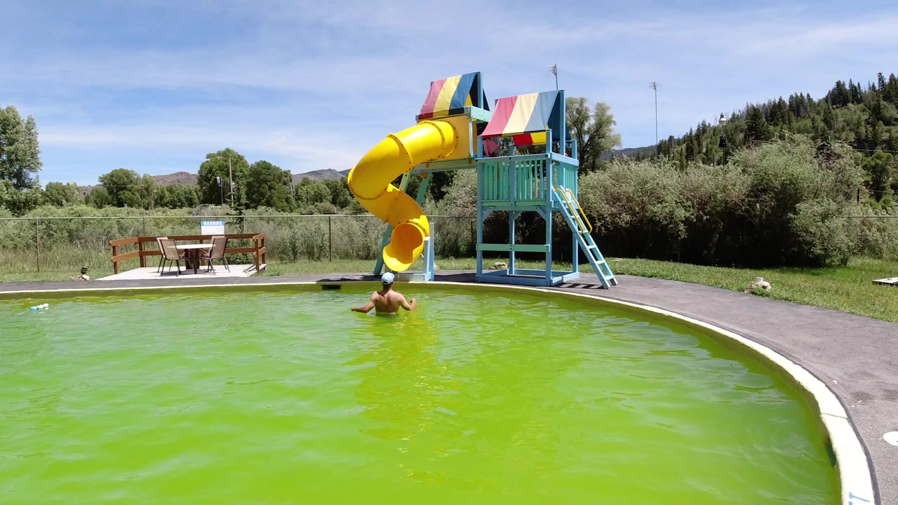 The wide frame shows the man slightly closer to the slide side of the green pool, with the yellow tube slide and blue stair structure prominent under bright daylight in Winter Park, Colorado.
