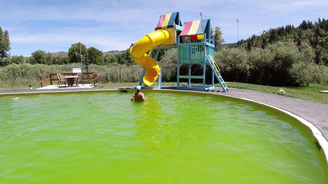 Winter Park, Colorado frame with a swimmer near the foreground edge of green pool water, the yellow slide and blue play structure centered against trees and hills.