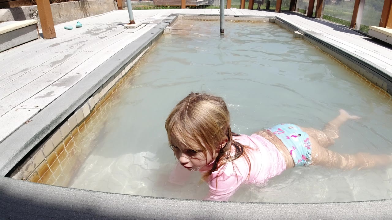 Young girl continues floating forward in the Winter Park, Colorado pool, with her face turned toward camera, legs extended behind her, and sunlit deck boards along the waterline.