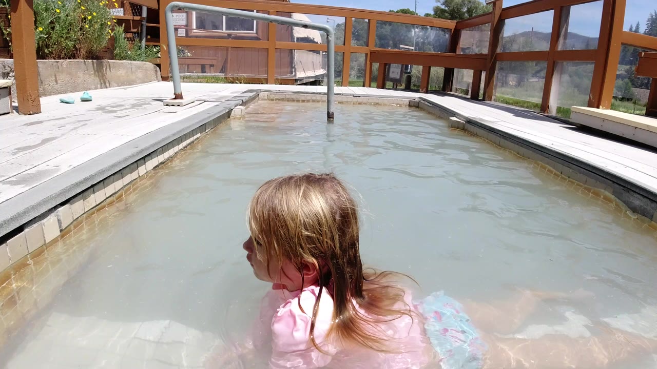 Young blond girl in a small outdoor pool beside a wooden deck railing.
