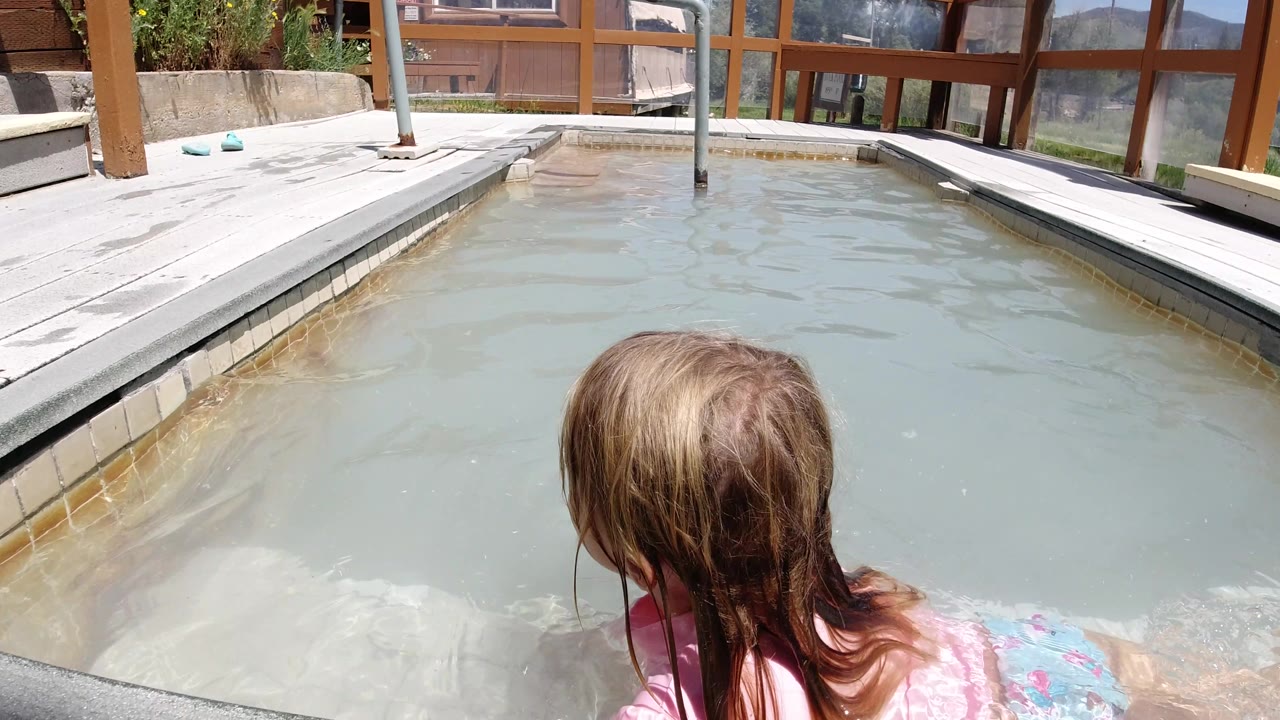 At 0:02, the child is seen more from behind in the Winter Park pool, with wet hair centered against pale water and a sunlit deck, windows, and wooden structure across the top of frame.