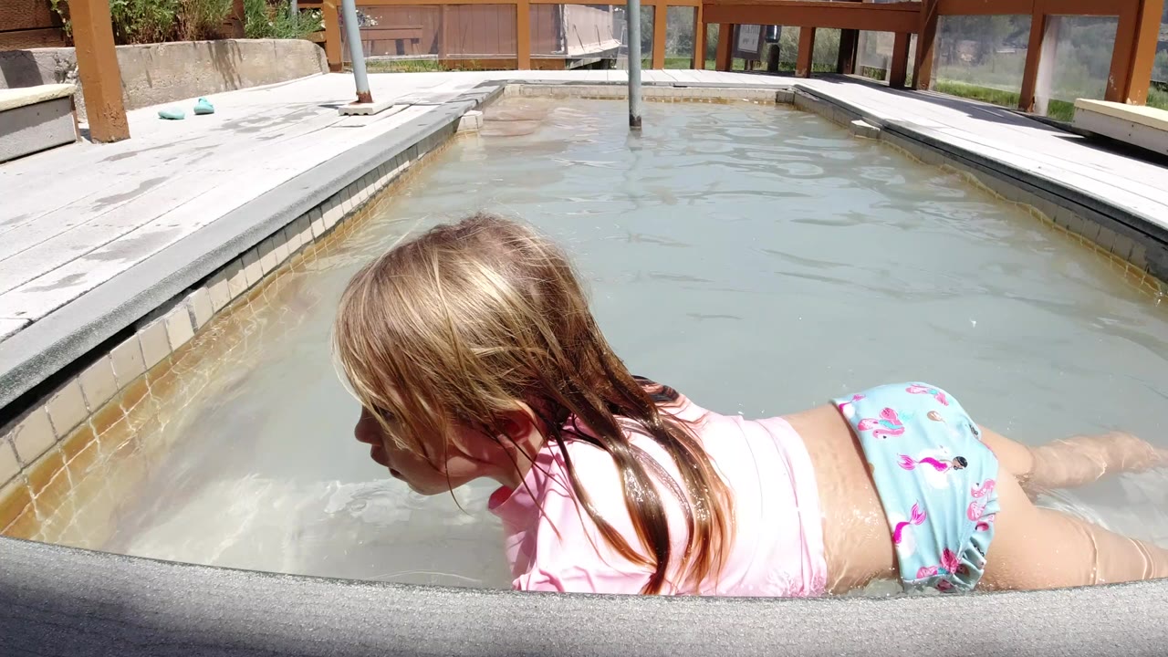 At 0:00 in Winter Park, Colorado, a young girl in pink and blue swimwear leans forward in cloudy pool water beside a deck and wood-framed glass wall.