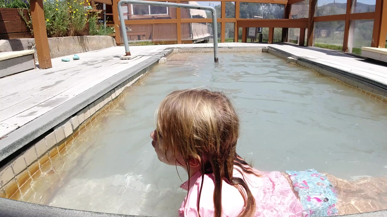 Young girl in a pink swim shirt lies partly in cloudy pool water at a home setting in Winter Park, Colorado, with deck boards, wooden posts, and windows behind her.