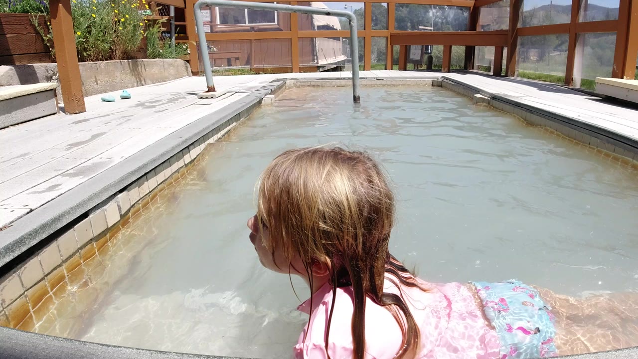 At one second, the girl remains near the near edge of the small pool in Winter Park, Colorado, looking left as sunlight hits her wet hair and the pale deck around the water.