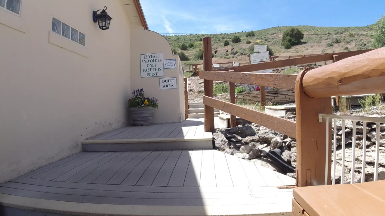 A pale exterior wall and wooden railing frame a sunny deck area in Winter Park, Colorado, with scrub-covered hills in the background and no visible people.