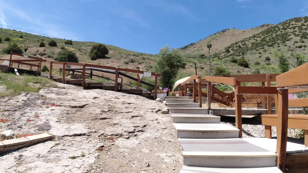 Stone steps rise toward brown wooden railings in Winter Park, Colorado, with rocky foreground, green hillside grass, blue sky, and no visible people.