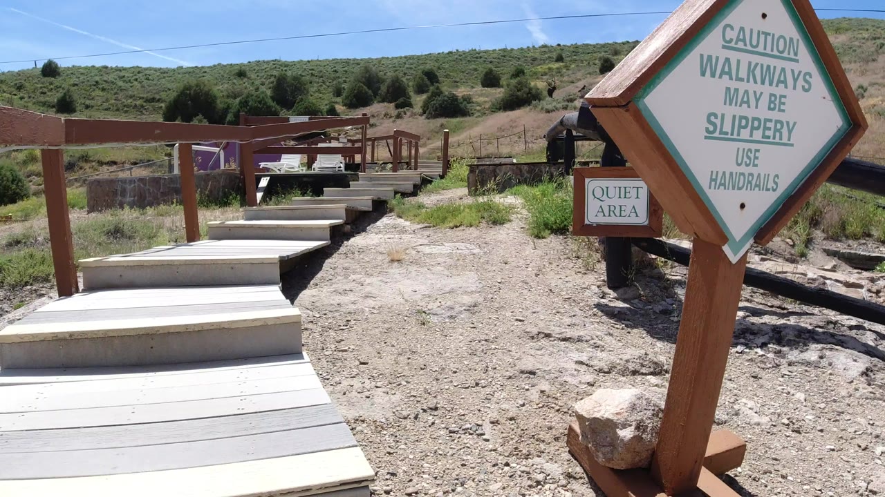 Wooden walkway steps and a large caution sign stand in a dry outdoor area in Winter Park, Colorado, with scrubby hills and no visible people.