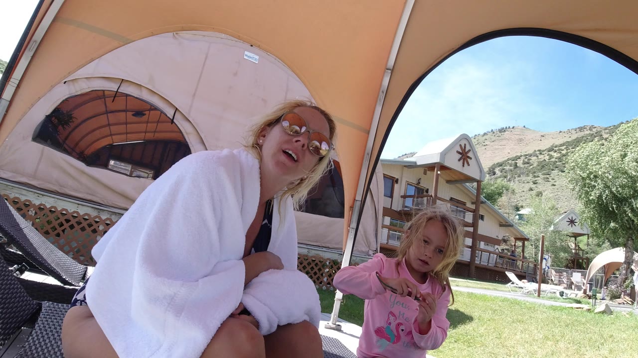 In Winter Park, Colorado, a woman wrapped in a white towel sits close to the camera while a young girl stands beside her under a tan canopy, with grass and lodge buildings behind them.