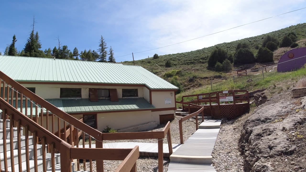 Winter Park, Colorado frame showing a light building with a pale green roof beside wood railings, gravel paths, rocky ground, hillside shrubs, and no visible people.