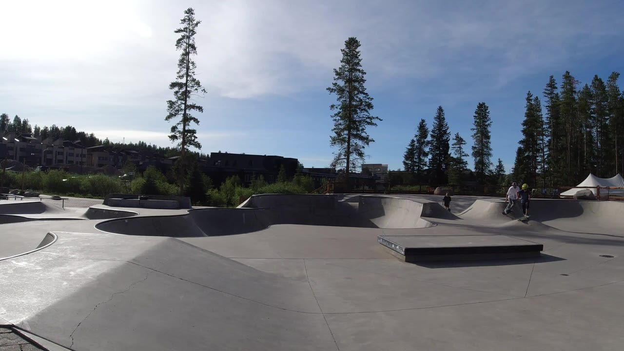 Wide concrete skate park with ramps, trees, distant buildings, and people near the bowls.