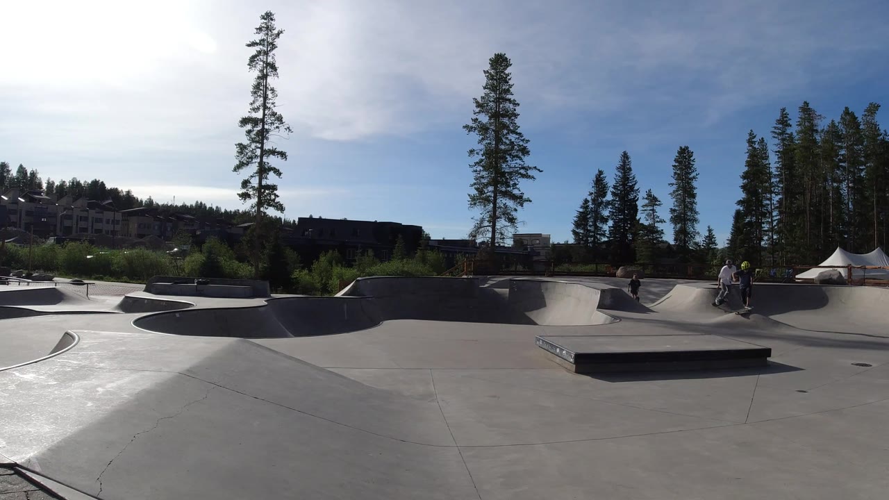 Wide Winter Park, Colorado skate park view with a person on or near a skateboard in the middle distance, a white tent at right, trees, buildings, and hard sun from the left.