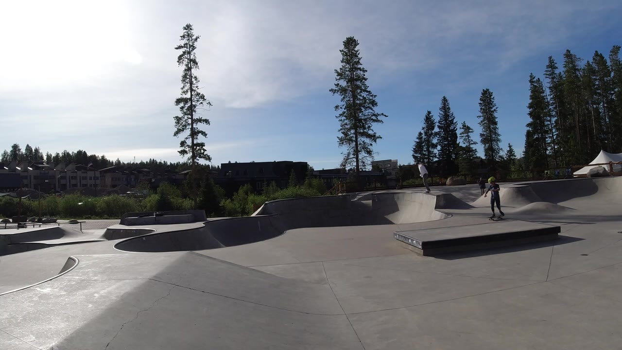 Wide view of a Winter Park, Colorado skatepark with a young skateboarder near a central ledge, concrete bowls, pine trees, distant buildings, and bright sun glare.