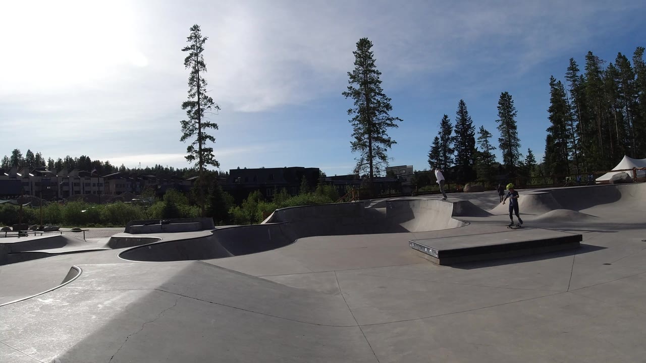 A young skateboarder stands near the rectangular ledge in the Winter Park skatepark, with curved ramps, tree line, distant buildings, and high backlit sky visible.
