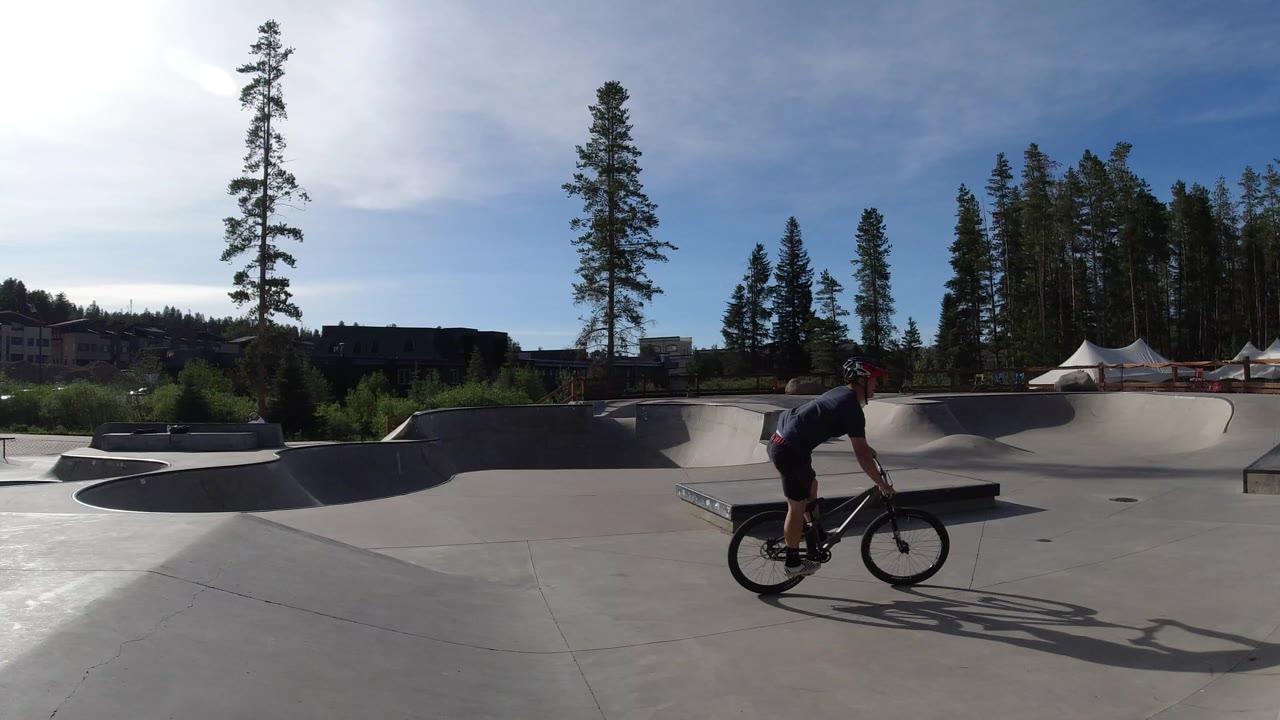 Cyclist rides a bicycle across a concrete skatepark under bright sun.