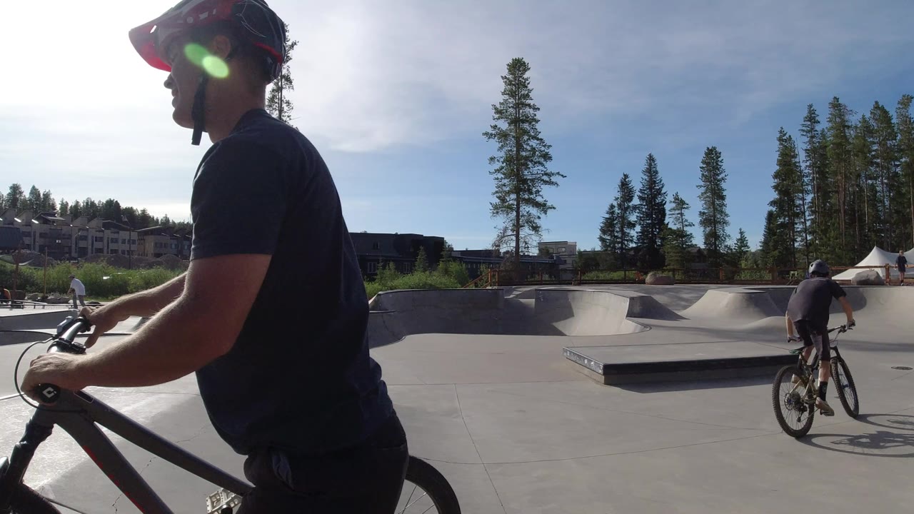 A foreground rider in a helmet occupies the left side while a second cyclist rides into the Winter Park concrete track, with pine trees and white tents beyond the ramps.