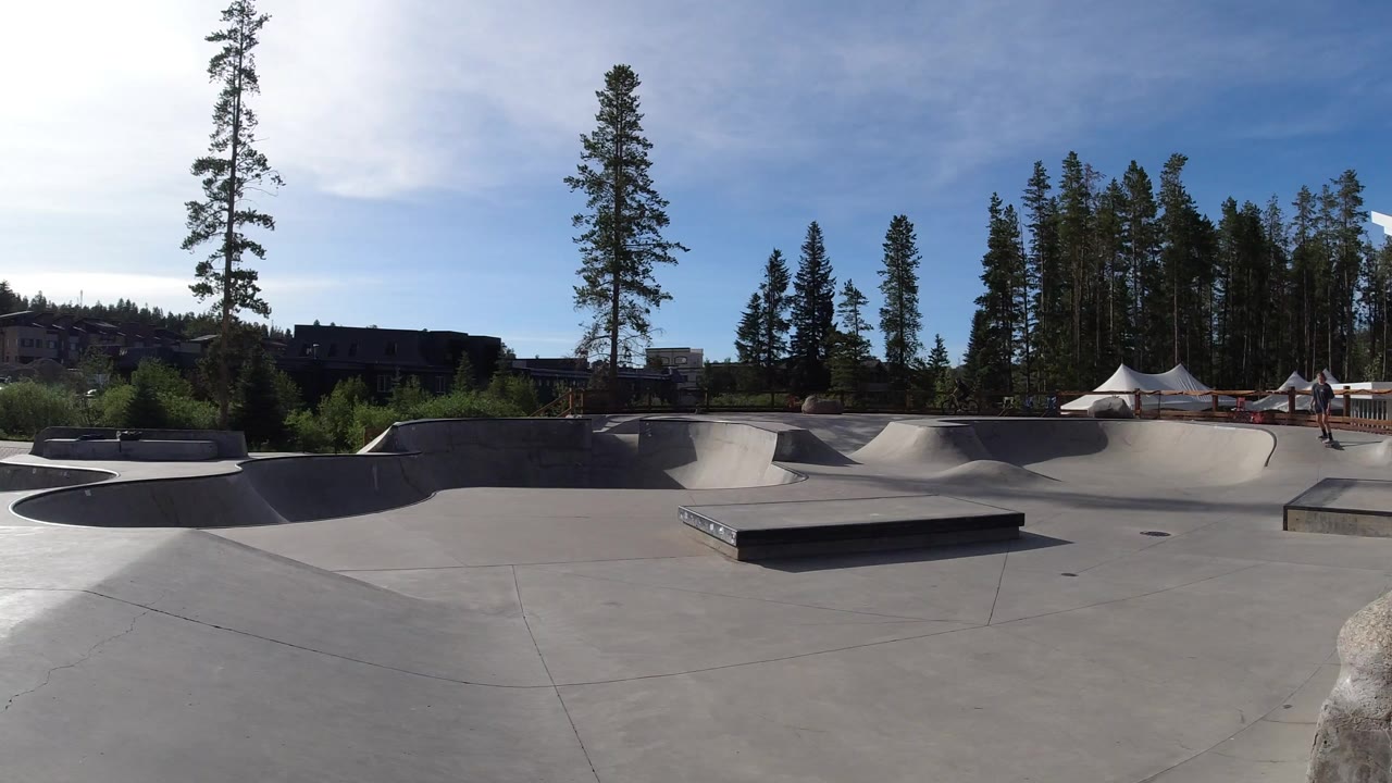 Wide concrete skatepark with ramps, trees, tents, and no visible people in the foreground.