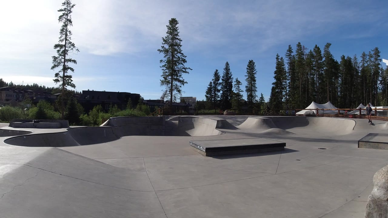 Wide Winter Park, Colorado skatepark frame with concrete bowls and a central ledge, tall trees behind it, white tents at right, and a small visible person near the far-right edge.