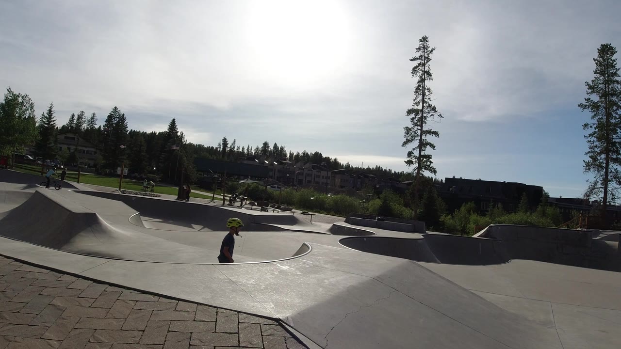 Wide view of a Winter Park, Colorado skate park with a helmeted rider near the concrete bowl, pine trees, buildings, and bright sun overhead.