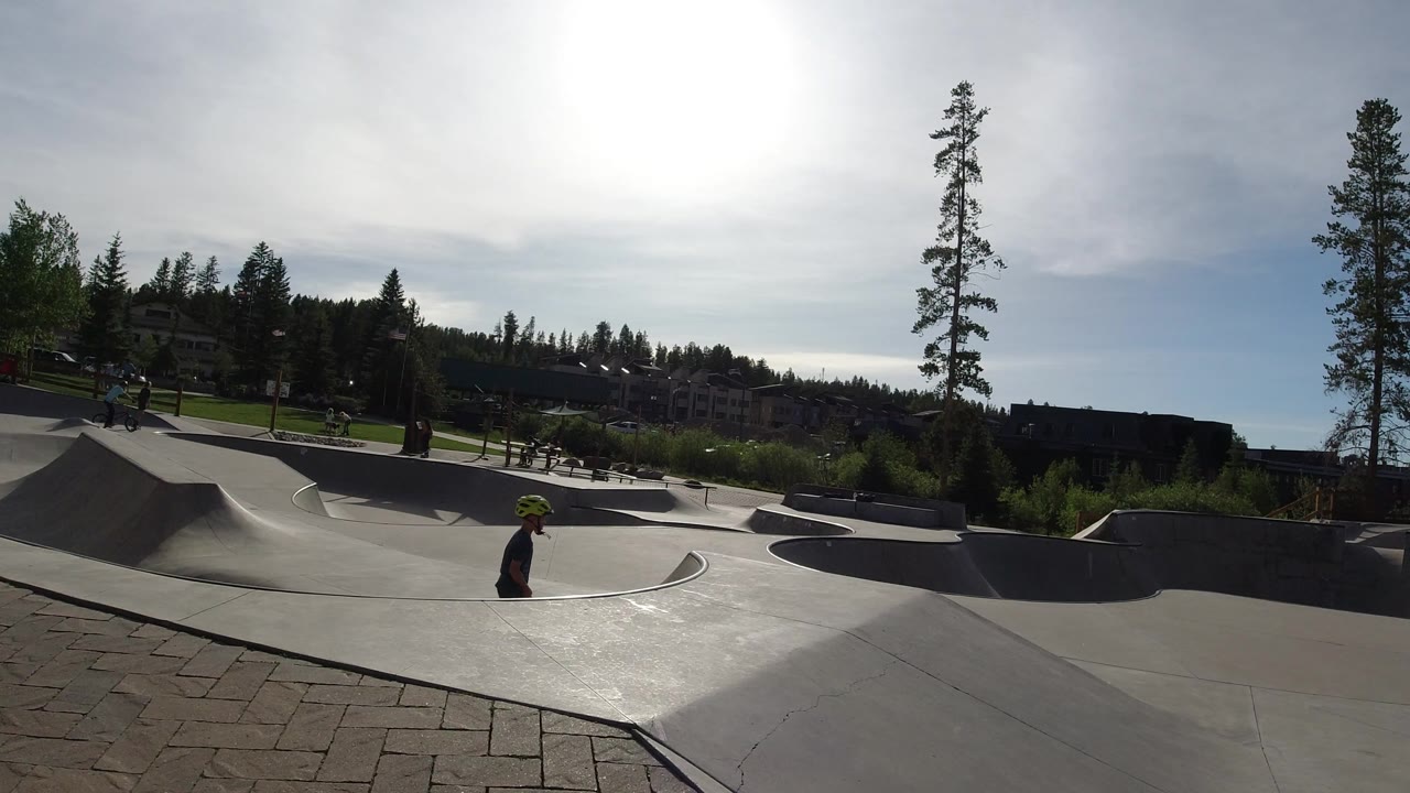 Winter Park skate park frame with the helmeted rider lower in the bowl, several distant park users, concrete ramps, pine trees, and strong overhead light.
