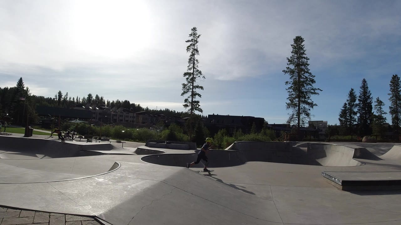Wide view of a Winter Park, Colorado skate park with concrete bowls, a low box, tall evergreens, hillside buildings, bright sun glare, and distant people skating.