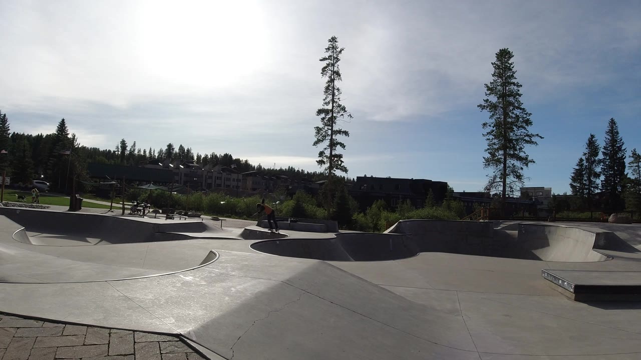 Wide view of a concrete skate park in Winter Park, Colorado, with ramps, pine trees, distant buildings, bright sun glare, and small distant people visible.