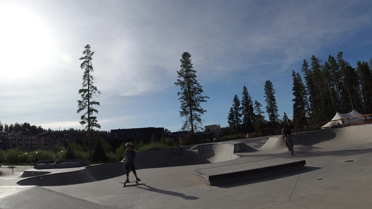 Wide concrete bike park with a rider crossing at left and another cyclist near the ramps.