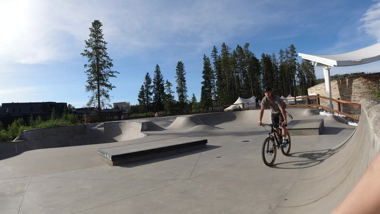 The rider moves closer through the Winter Park concrete skatepark, framed by the low grind box on the left, curved ramps, sun glare, and tall trees behind him.