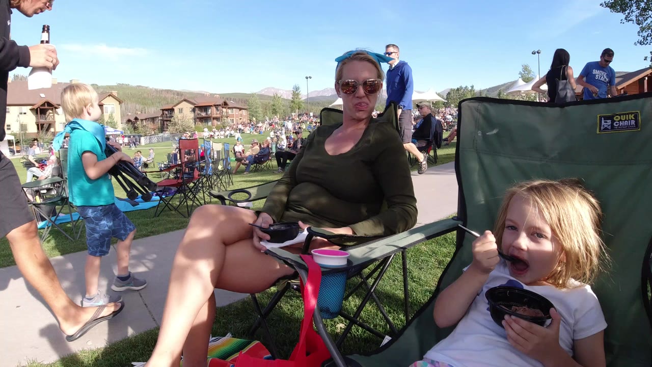 In Winter Park, Colorado, a woman in sunglasses sits beside a young child in folding chairs while people and a lawn crowd fill the background.